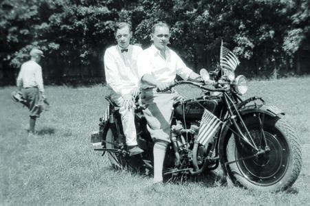 Black and white photograph of two men in collared shirts astride an Indian motorcycle in…